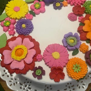 May include: A white cake decorated with colorful, edible flowers. The flowers are in various shapes and sizes, including yellow, pink, red, orange, and purple. The cake is on a white, lacy cake stand, creating a festive and celebratory appearance.