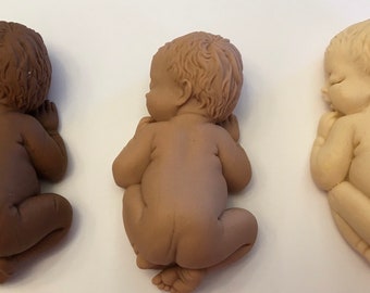 Può includere: Tre statuette di bambini che dormono in diverse tonalità di pelle: marrone scuro, marrone medio e beige chiaro. I bambini sono scolpiti in posizione fetale, con gli occhi chiusi e le mani vicino al viso. Le statuette sono probabilmente decorative.