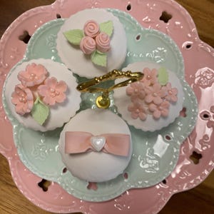 Puede incluir: Una bandeja de servicio de varios niveles con cuatro cupcakes, cada uno decorado con flores y lazos de color rosa pastel. Las magdalenas se encuentran en un plato verde claro, que está sobre un plato rosa más grande con volantes. Un asa dorada está en el centro.