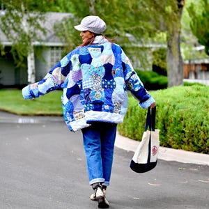 May include: A quilted jacket featuring a patchwork of various blue and white fabric patterns, worn with blue jeans, a grey newsboy cap, and a white canvas tote bag with dark blue handles and a circular graphic print on the side.