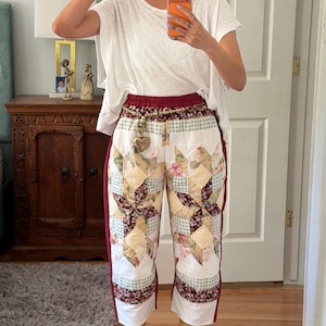 Può includere: Pantaloni patchwork con motivo floreale e geometrico, cintura e bordi laterali bordeaux. I pantaloni sono abbinati a stivali color cuoio e una maglietta bianca. Viene indossato un berretto da baseball con testo.