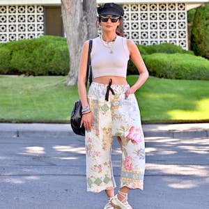 May include: A beige ribbed sleeveless crop top paired with wide-leg cropped pants featuring a multi-colored floral and geometric patchwork quilt pattern, a black newsboy cap, dark sunglasses, a black shoulder bag, and white lace-up flats.