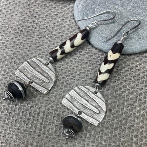 May include: A pair of dangle earrings featuring a silver-toned hook, a cylindrical bead with a black and cream patterned design, a textured silver-toned semi-circular metal component, and a small black bead with a silver-toned metal cap at the base.