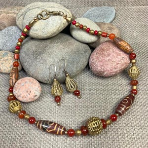May include: A necklace and earring set featuring a mix of red, gold, and brown beads. The necklace has a variety of bead shapes, including round, oval, and cylindrical. The earrings have a gold-colored, cage-like design with red beads.