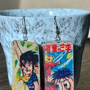 May include: Handmade earrings featuring vintage Japanese manga illustrations. The rectangular earrings display colorful comic book panels with detailed character art and text. The earrings hang from silver-colored ear wires.