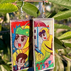 May include: Rectangular earrings featuring colorful anime-style illustrations. The earrings depict characters in yellow and red outfits with silver fish hook ear wires. The background includes green leaves.
