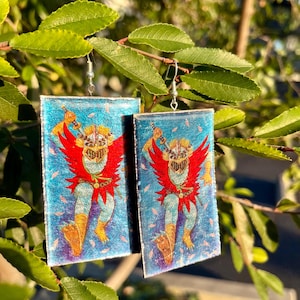 May include: A pair of rectangular earrings with a vibrant illustration of a figure with red wings, a mask, and a blue background. The earrings are suspended from silver-colored hooks, showcasing a unique and artistic design. The earrings are hanging from a leafy branch.
