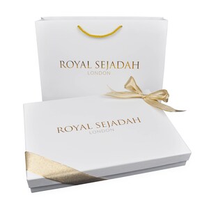 以下が含まれることがあります： 白いギフトボックスに金色のリボンがかけられており、上部に「ROYAL SEJADAH LONDON」の文字が書かれています。ボックスは、同じ文字が書かれた白いショッピングバッグの上に置かれています。