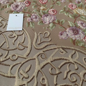 Op de afbeelding: Een beige gebedskleed met een bloemenpatroon en ingewikkelde kalligrafie in goud en bruin. Het kleed heeft de tekst "Royal Sejadah London" op een wit label.