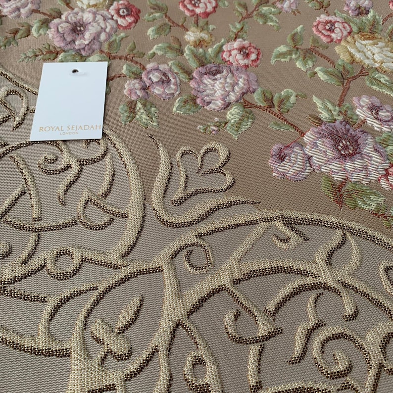 Op de afbeelding: Een beige gebedskleed met een bloemenpatroon en ingewikkelde kalligrafie in goud en bruin. Het kleed heeft de tekst "Royal Sejadah London" op een wit label.