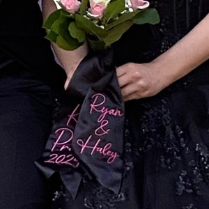 May include: A bouquet of white and pink roses tied with a black satin ribbon with the text "Ryan & Haley 2021" in pink.