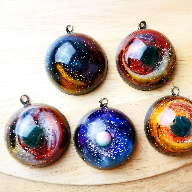 Pu&ograve; includere: Cinque ciondoli galassia fatti a mano con anelli color argento. Ogni ciondolo presenta un design a spirale unico con colori come blu, rosso, arancione e bianco, che ricordano nebulose e galassie. I ciondoli sono rotondi e hanno una finitura lucida.