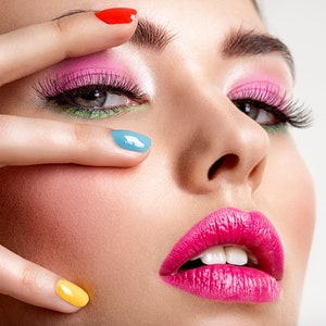 Pu&ograve; includere: Primo piano di un viso con trucco audace. Ombretto rosa e accenti verdi evidenziano gli occhi, con lunghe ciglia. Le labbra sono di un fucsia brillante e le unghie sono dipinte di rosso, blu e giallo. La pelle ha un incarnato rosato.