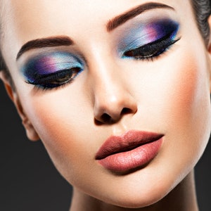 Pu&ograve; includere: Primo piano di un viso con un trucco occhi drammatico. L'ombretto presenta una miscela di tonalit&agrave; blu, viola e rosa. Le labbra sono di una tonalit&agrave; rosa naturale. La pelle ha un aspetto liscio e impeccabile.