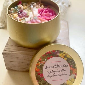 May include: A gold-colored candle tin with a floral design. The candle is filled with wax and decorated with pink rose petals and other dried flowers. The lid features a floral design and the text "Secret Garden".