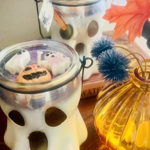 May include: Two white glass ghost-shaped candle jars with black painted eyes and mouths. The candles contain wax embeds of small white ghosts and an orange pumpkin. Each jar has a black wire handle and a small tag with a spider illustration.