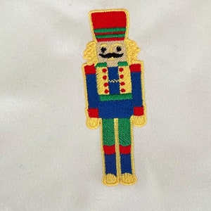 Op de afbeelding: Witte linnen servet met een geborduurd notenkrakerontwerp. De notenkraker draagt ​​een rode hoed, een blauw jasje en een groene broek.