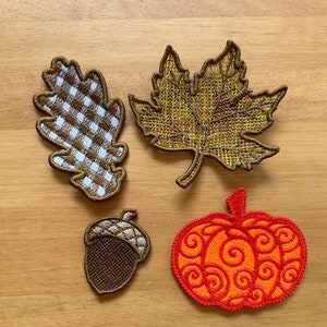 Pode incluir: Quatro patches bordados com designs outonais: uma folha de carvalho xadrez marrom e branco, uma folha de bordo amarela, uma bolota marrom e uma abóbora vermelha com um padrão de espiral.
