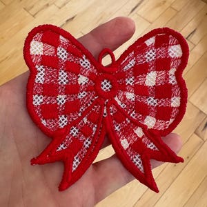 May include: A red and white gingham bow-shaped embroidered applique. The bow has a loop for hanging and is held in a hand. The bow is red with white accents and a white border.