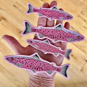Op de afbeelding: Vier roze zalmvis-geborduurde patches met groene vinnen en zwarte ogen. De patches zijn gerangschikt in aflopende volgorde van grootte, met de grootste patch bovenaan en de kleinste patch onderaan.
