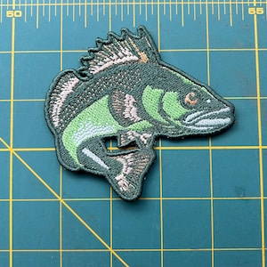 May include: A green and white embroidered patch of a fish with a fin on its back. The fish has a light orange eye and a white mouth.