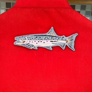 Könnte beinhalten: Ein roter Stoff-Patch mit einem gestickten Bild einer Forelle. Der Fisch ist grau und weiß mit schwarzen Flecken. Der Fisch schaut nach rechts.