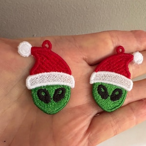 Könnte beinhalten: Zwei gestickte Aufnäher mit grünen Alienköpfen, die rote Weihnachtsmützen mit weißem Rand und einem weißen Bommel tragen. Die Aufnäher haben schwarze ovale Augen und eine kleine Schlaufe zum Aufhängen.