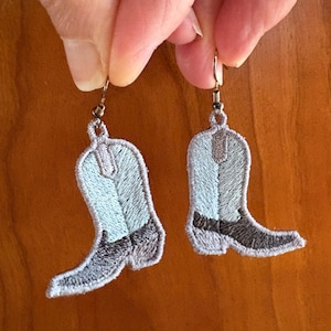 Puede incluir: Pendientes bordados de bota de vaquero. Los pendientes presentan un esquema de color azul claro y gris con detalles en marrón oscuro. Las botas están detalladas con costuras y cuelgan de ganchos plateados.