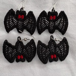 May include: Four pairs of black lace bat earrings with silver-colored hooks. Each bat has red accents on the face. The earrings are displayed on a white surface.