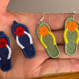 以下が含まれることがあります： 刺繍されたビーチサンダルのイヤリングとアップリケ。イヤリングは、黄色のストラップとオレンジ色の花が付いた緑色です。アップリケは、白いストラップと赤い花が付いた青色です。イヤリングにはシルバーのフックが付いています。