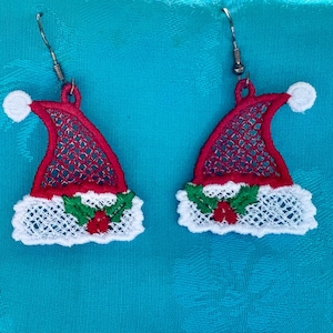 May include: A pair of festive Christmas earrings. Each earring features a red Santa hat design with a white pom-pom, a white lace trim, and a sprig of holly with red berries. The earrings have silver-colored fish hook ear wires.