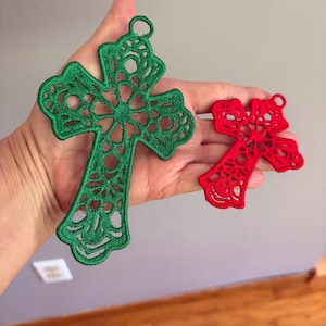 Könnte beinhalten: Zwei bestickte Kreuzornamente. Eines ist grün, das andere rot. Beide haben ein filigranes, durchbrochenes Design und eine kleine Schlaufe zum Aufhängen. Diese dekorativen Kreuze eignen sich für religiöse oder festliche Bastelarbeiten.