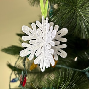 May include: White snowflake ornament hanging on a green Christmas tree with a green ribbon.