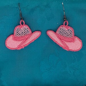 Könnte beinhalten: Ein Paar rosa bestickte Cowboyhut-Ohrringe. Die Ohrringe haben ein detailliertes Design mit einem Netz-Kronenteil und einer gebogenen Krempe. Die Ohrringe sind an silberfarbenen Fischhaken-Ohrdrähten befestigt.