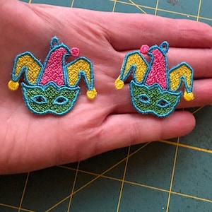May include: Two embroidered patches in the shape of a jester's hat and mask. The patches feature a pink hat, yellow and blue accents, and a green mask with eye holes. The patches are held in a hand.