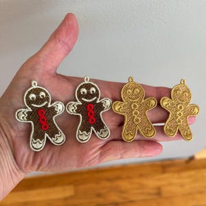 May include: Four gingerbread man ornaments. Two are brown with white and red accents, and two are gold. Each ornament has a loop at the top for hanging. The gingerbread men have smiling faces and are suitable for holiday decor.