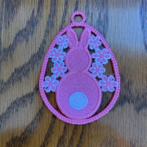 May include: Pink Easter egg ornament with a white bunny inside and white flowers around the bunny. The ornament has a loop at the top for hanging.