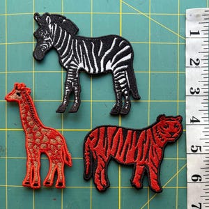 May include: Three embroidered animal patches: a zebra with black and white stripes, a giraffe with orange and brown spots, and a tiger with red and black stripes. The patches are on a green surface, with a ruler showing inches.