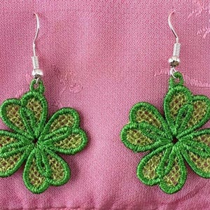 May include: A pair of green four-leaf clover earrings. The earrings are made of embroidered fabric with a silver-colored hook. The clover design has a darker green outline and a lighter green center. The earrings are displayed on a pink fabric.