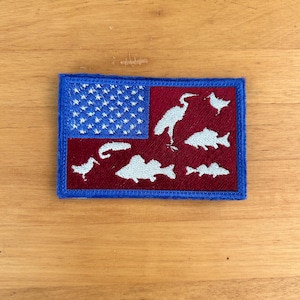 May include: A blue and red embroidered patch with a white silhouette of a heron and fish on a red background. The top left corner of the patch features a blue and white American flag.