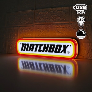 Puede incluir: Un letrero de neón rojo, amarillo y blanco con la palabra "MATCHBOX" en letras negras. El letrero está iluminado y tiene un reflejo en una superficie oscura. El letrero se alimenta mediante un cable USB y tiene un interruptor de atenuación.