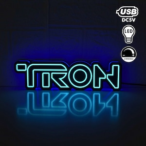 Puede incluir: Un letrero de neón azul con la palabra "TRON" en letras negras delineadas. El letrero está iluminado y tiene un reflejo en una superficie negra. El letrero se alimenta mediante un cable USB y tiene un interruptor de intensidad.
