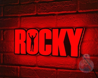 Puede incluir: Señal iluminada roja con la palabra "ROCKY" en letras negras. Una silueta de una persona con los brazos levantados está en el medio de la palabra "ROCKY".
