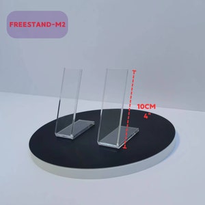 Peut inclure: Deux présentoirs en acrylique transparent, mesurant chacun 10 cm de haut, sont présentés sur une base circulaire noire. Les présentoirs sont conçus pour maintenir les objets à la verticale.