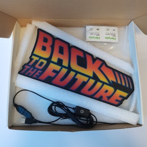 Op de afbeelding: Een zwart en rood bord met de tekst "BACK TO THE FUTURE" in een gestileerd lettertype. Het bord zit in een doos met schuimvulling, een USB-kabel en reinigingsdoekjes.