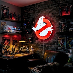 May include: A room decorated with Ghostbusters memorabilia. A large, illuminated sign of the Ghostbusters logo is mounted on a brick wall. Shelves display action figures, posters, and a model of the Ecto-1 car. A desk holds more figures.