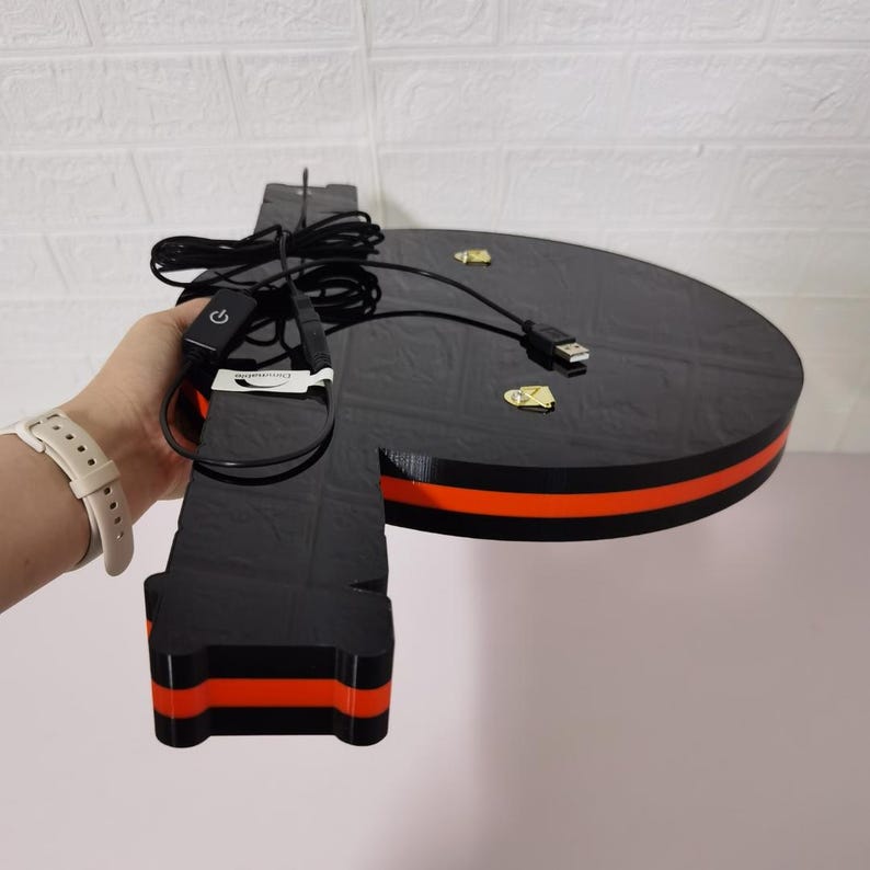 K&ouml;nnte beinhalten: Schwarzes und oranges kreisf&ouml;rmiges Objekt mit einem Ein-/Ausschalter und einem USB-Kabel. Das Objekt hat eine schwarze Oberfl&auml;che mit einem orangefarbenen Streifen am Rand. Das Objekt wird von einer Person mit einer wei&szlig;en Uhr gehalten.