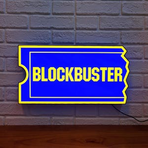 May include: A rectangular sign with the word "BLOCKBUSTER" in yellow letters on a blue background. The sign has a yellow border and is mounted on a brick wall. A black cord extends from the sign.