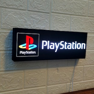 May include: A black rectangular sign with white LED lights. The sign reads "PlayStation" in white letters and features the PlayStation logo in the top left corner. The logo is a stylised blue, red and white "P" inside a circle.