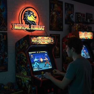 Könnte beinhalten: Eine Person spielt ein Mortal Kombat Arcade-Spiel. Der Spielautomat ist schwarz und rot mit dem Logo des Spiels. Ein Neonschild mit dem Mortal Kombat-Logo und einem Drachen ist an der Wand angebracht. Poster und andere Arcade-Spiele sind im Hintergrund zu sehen.
